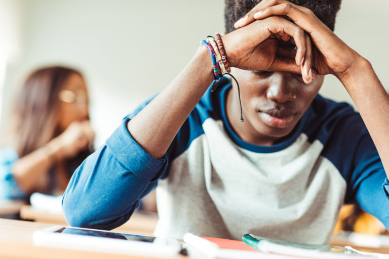 Male student resting face on hands, sitting at his desk, female student blurred in background