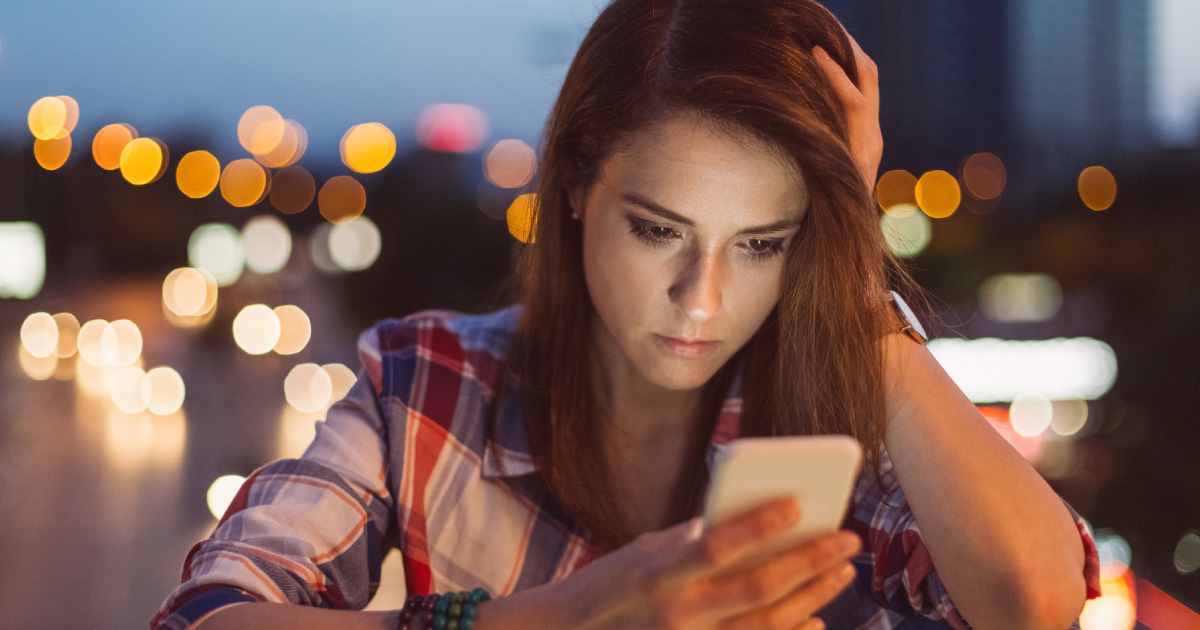 Young woman looks at phone screen, one hand on head, background lights blurred into orbs