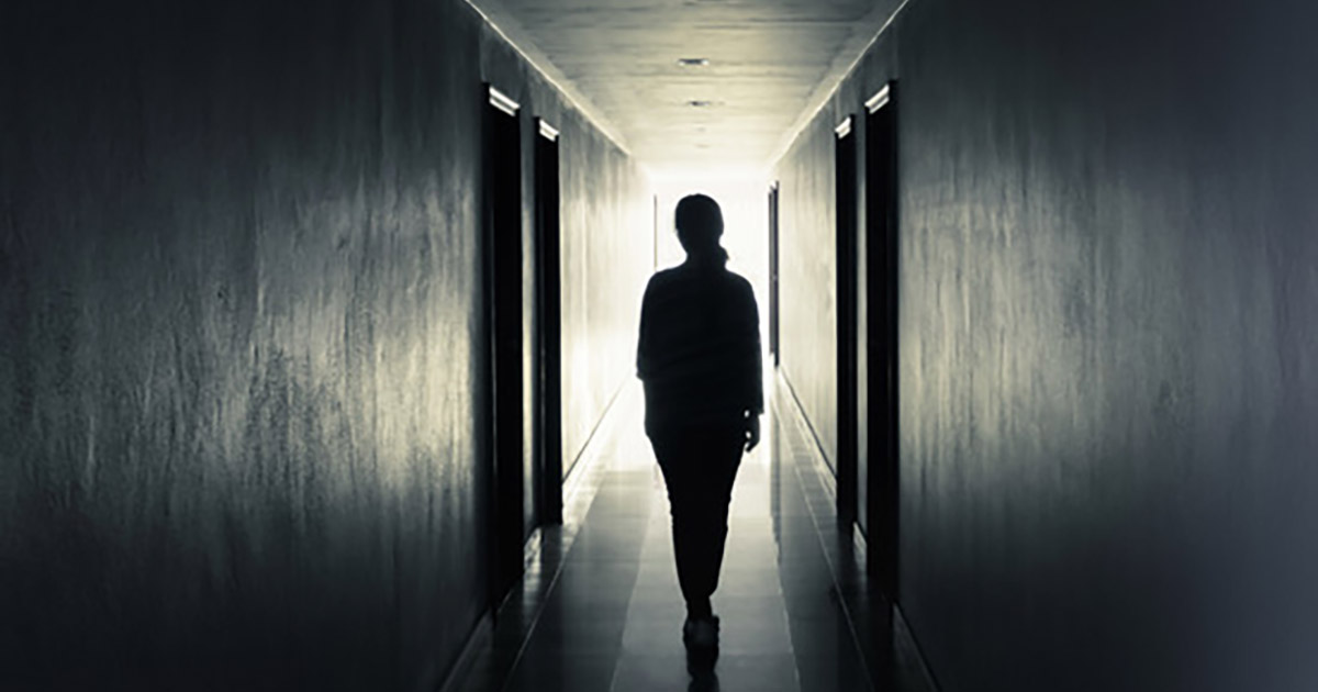 Woman walks down hallway with doors on either side, hallways is lit at the end, woman silhouetted by light