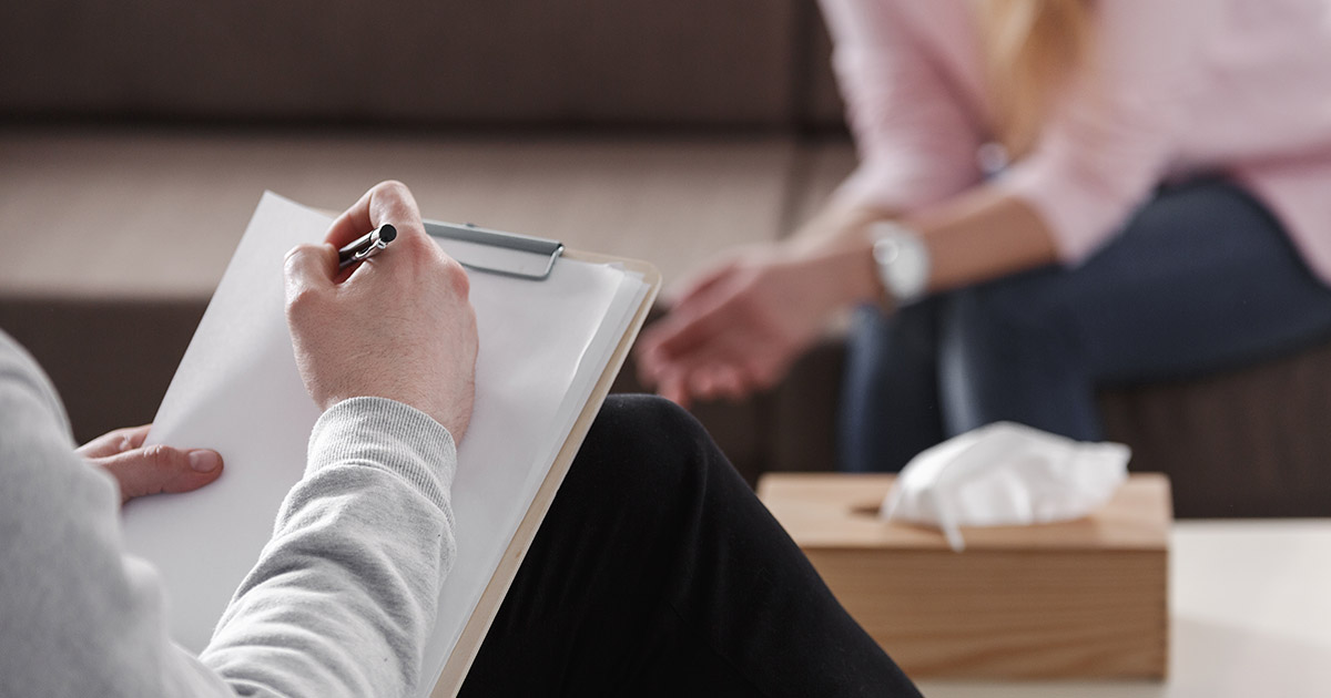 Man holds clipboard and pen, seated facing a woman, partially in view, hands clasped at knees, facial tissue and wooden holder between them