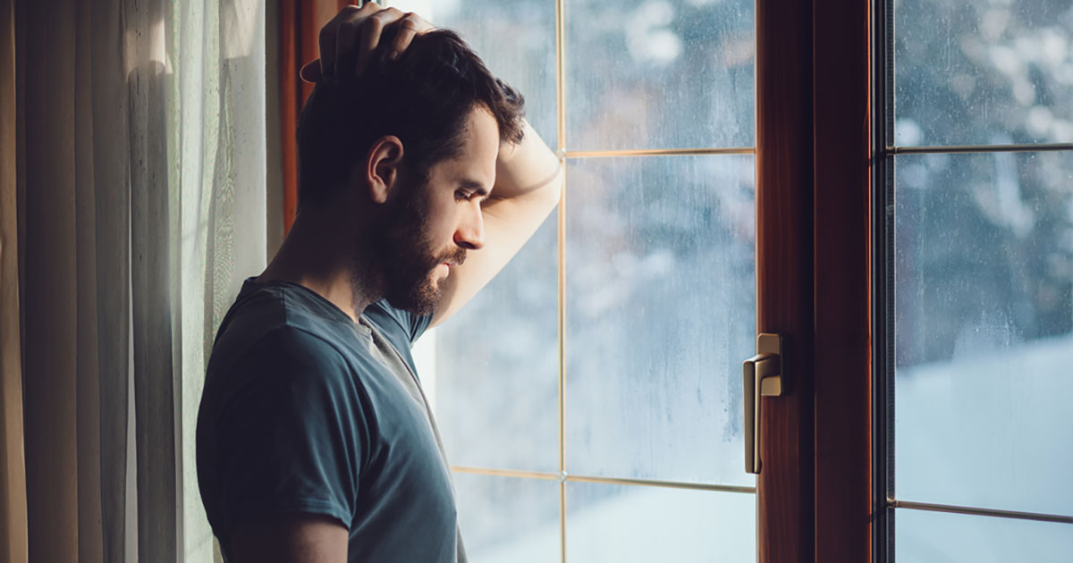 man in short sleeve shirt stands at paned window, looking pensive, elbow resting on glass and hand in hair, window streaked with rain