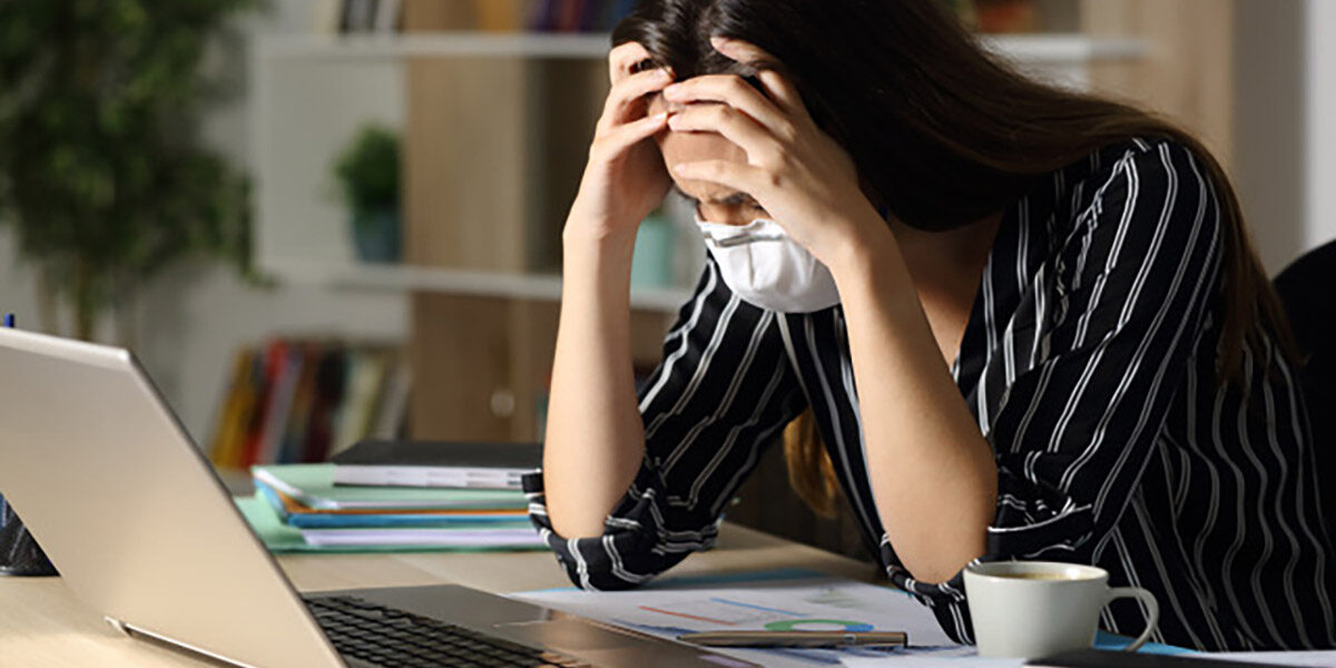 Woman wearing button up shirt, sleeves rolled up, holds head, mask on face, seated at desk looking pensive