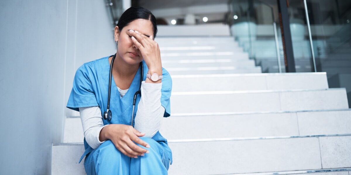 Health care worker seated, squeezing bridge of nose, eyes closed, looking stressed.