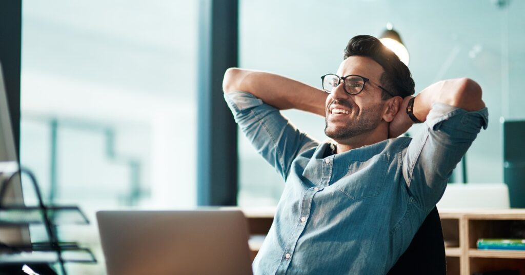 A man sitting at an office desk, leaning back in his chair with his hands behind his head. He looks upward and smiles.