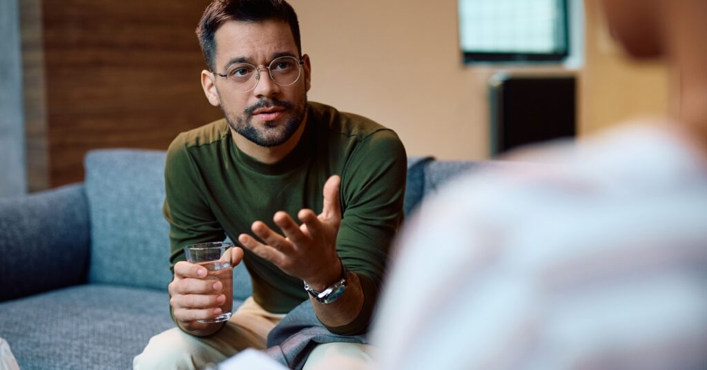 A seated man talks with a neutral expression and gestures with an open hand. The person in the foreground holds a clipboard.