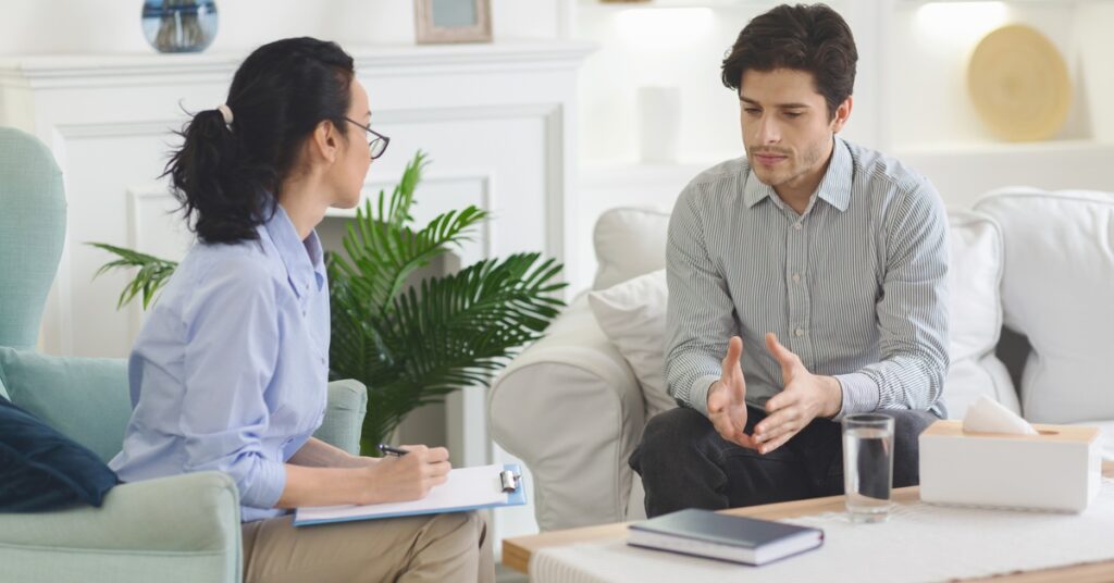 A man sits on the edge of a couch and talks to a therapist. He gestures with his hands near his waist and has downcast eyes.