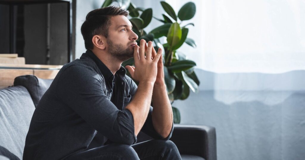 Side view of a man sitting on a couch near a window. He leans forward with his elbows on his knees and gazes upward.