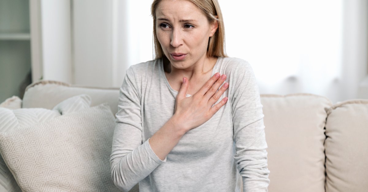 A woman sits up straight on the edge of a couch. She holds her hand over her heart and breathes through her mouth.