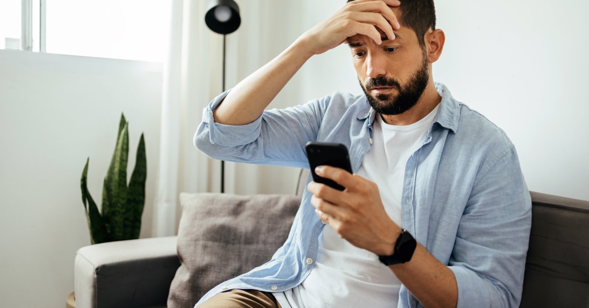 A man in a bright room looks at his smartphone screen with a furrowed brow. He holds his right hand over his forehead.