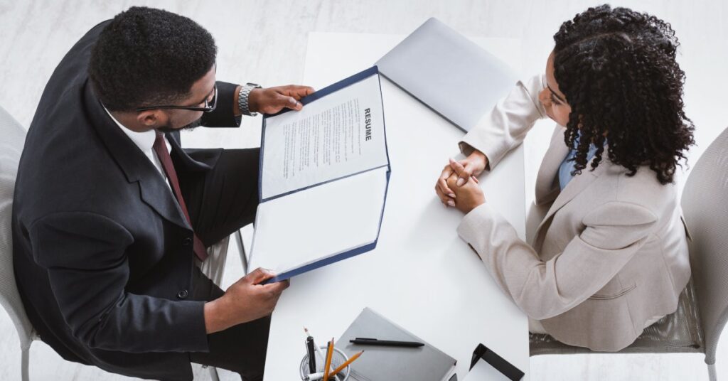 Overhead view of two professionals sitting at a desk. One holds open a folder and looks at a sheet titled "Resume."
