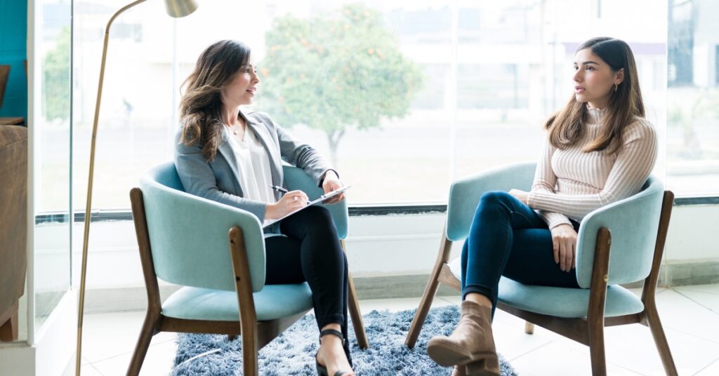 Two women sit in armchairs and talk. One writes on a clipboard, observing, while the other talks without meeting her gaze.