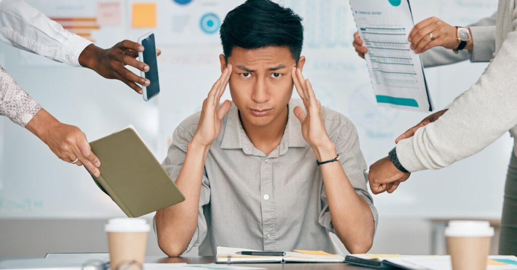 A man at a conference table rubs his temples. People around him show him a book, a phone, documents, and a watch.