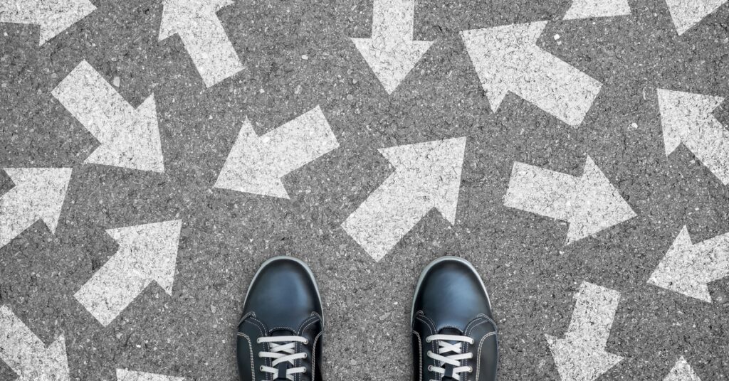 Downward view of someone standing in black shoes on asphalt concrete. White arrows on the ground point in different directions.