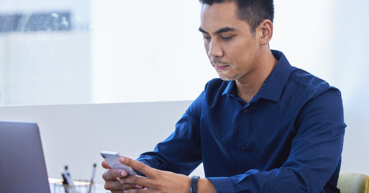 A man sits at a desk near a large window and looks at his cell phone. His laptop and an open notebook are on the desk.
