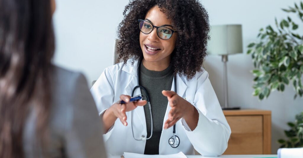 A doctor smiles and gestures as she speaks with a patient. Papers on a clipboard lie on the desk between them.