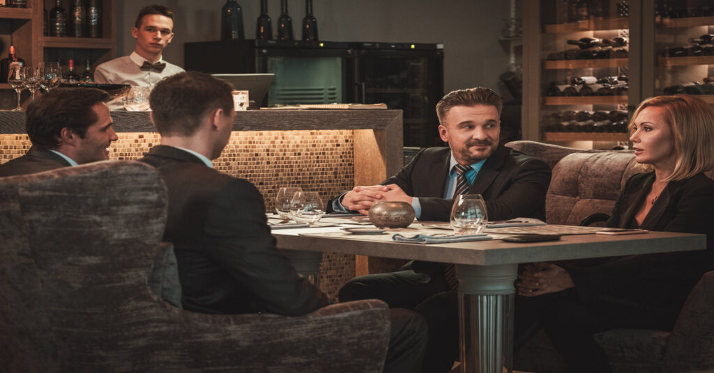 Four people wearing suits sit around a dinner table in front of a bar. Empty, stemless wine glasses are on the table.