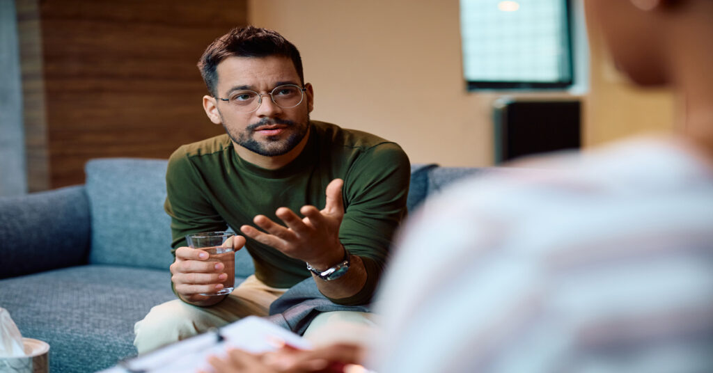 A man leans forward on a couch and talks to someone who has a clipboard on their lap. He holds a glass of water.