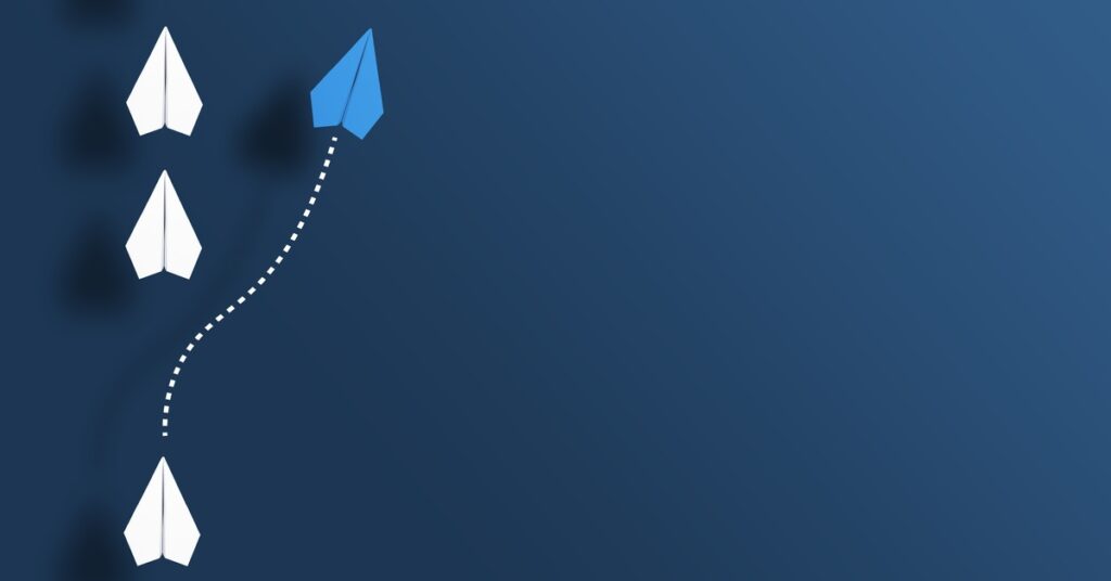 White paper airplanes are arranged in a line. One blue paper plane leaves the formation and glides to the right.