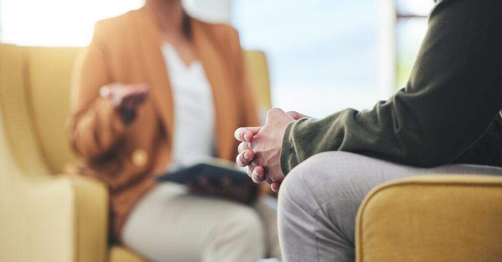 In the foreground, a man sits with his hands clasped tightly. In the blurred background, a woman holding an open book gestures.
