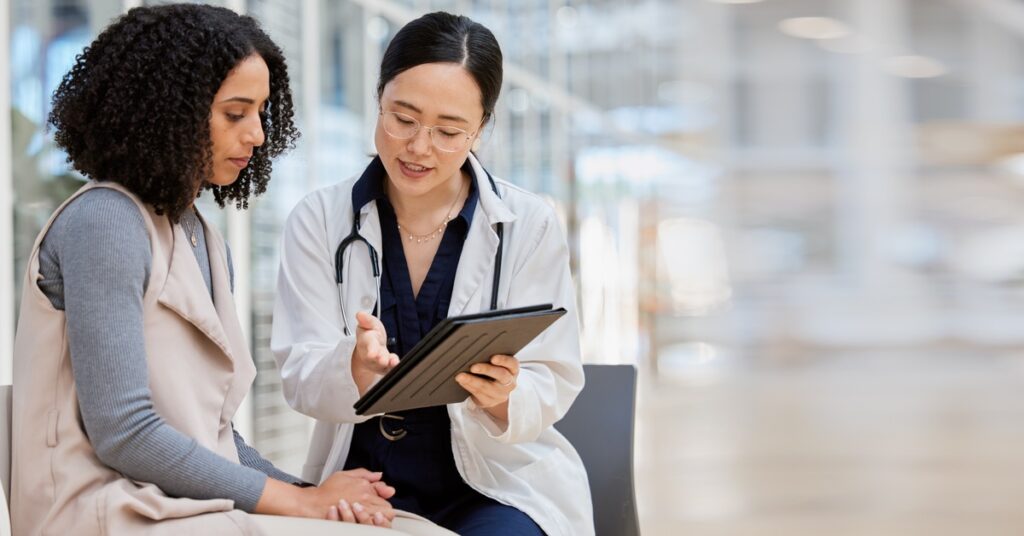 A patient looks at a digital tablet that their doctor shows them. The doctor talks and gestures at something on the screen.