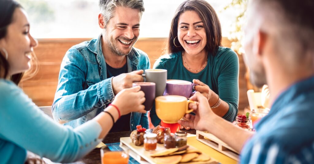 A group of smiling people toasts with their coffee mugs on a sunny day. Their table is covered with snacks or appetizers.