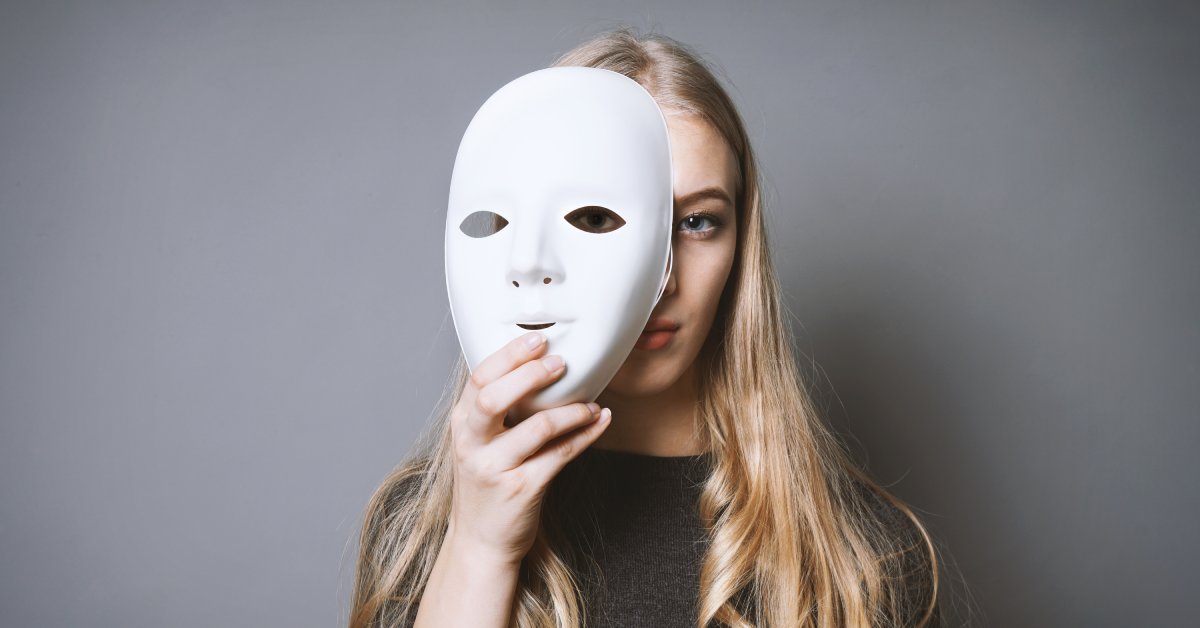 A woman holds up a white, full-face mask and partially covers her face with it. Her real face has a neutral expression.