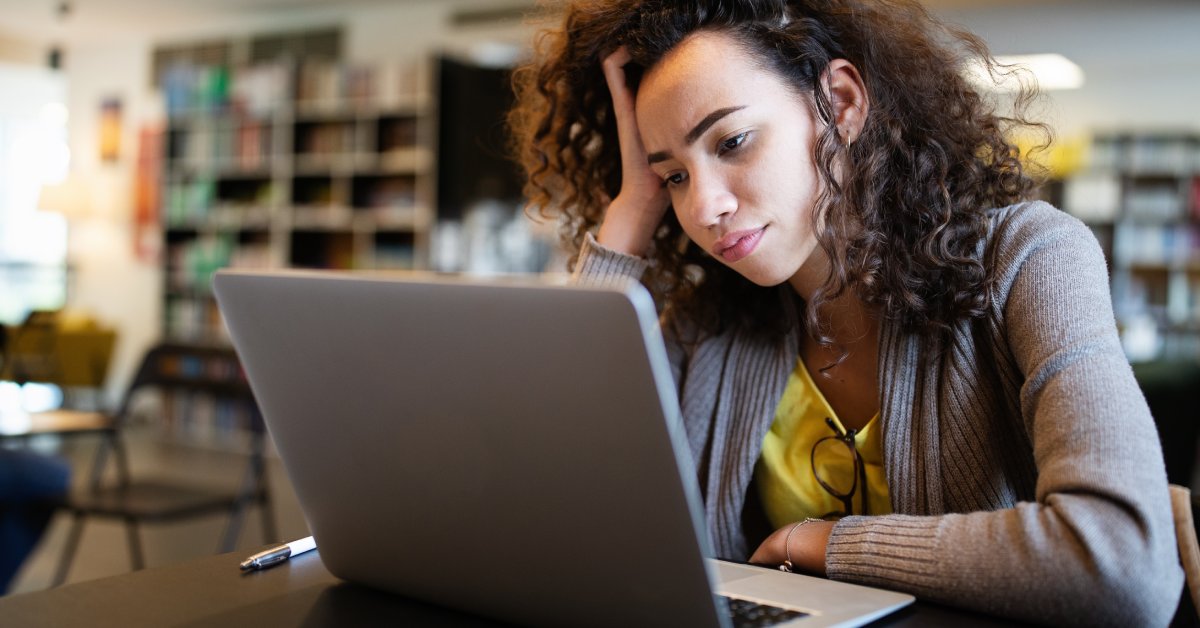 A woman sits at a table in a library. She props her head up with her hand and slightly frowns as she looks at a laptop screen.