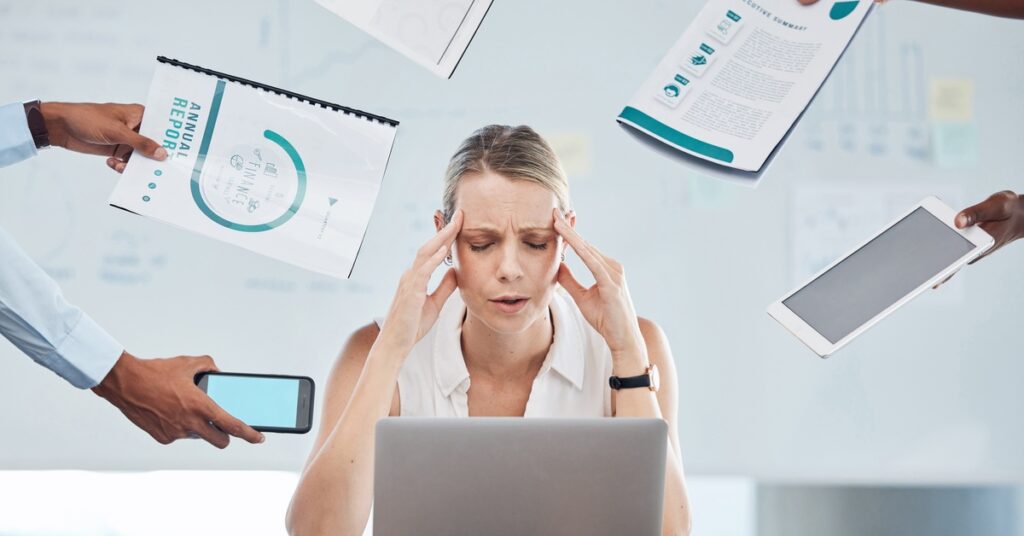 An office worker sits with closed eyes and rubs her head. People hold out papers, a digital tablet, and a cell phone to her.