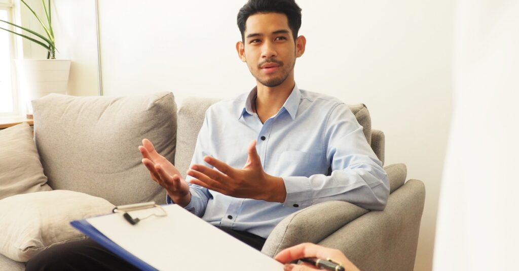 A young man in a sunny room sits on a couch and talks to someone holding a clipboard. He gestures with his palms face up.
