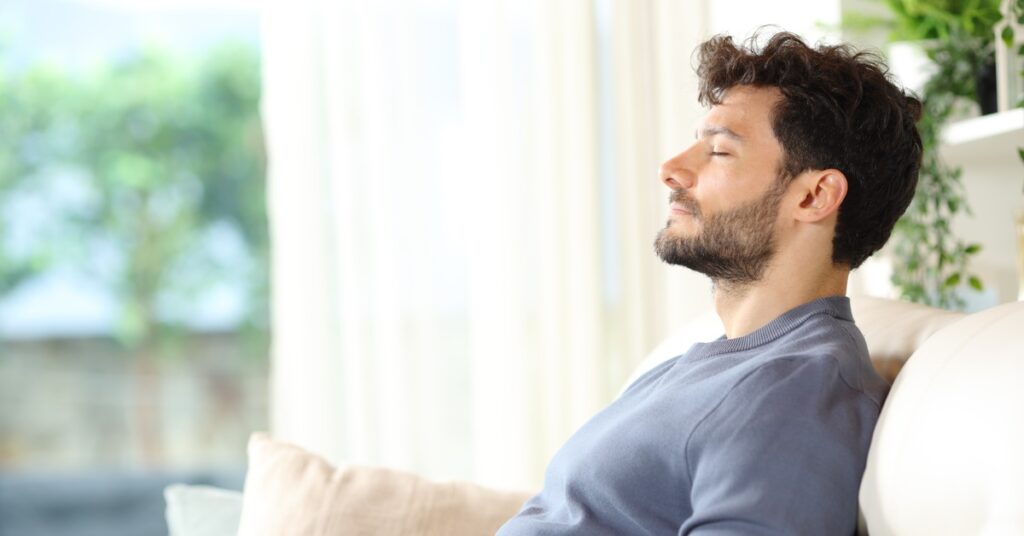 Side view of a man seated on a couch in a bright white room. He leans back with closed eyes and relaxed shoulders.