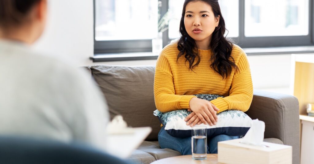 A woman rests her arms on a pillow on her lap as she talks to someone holding a clipboard. Water and tissues are on a table.