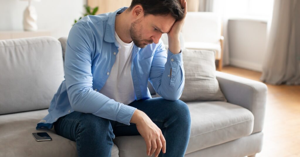 A man leans forward on a couch in a neutral-colored room. He presses his palm to his temple and frowns with his eyes open.