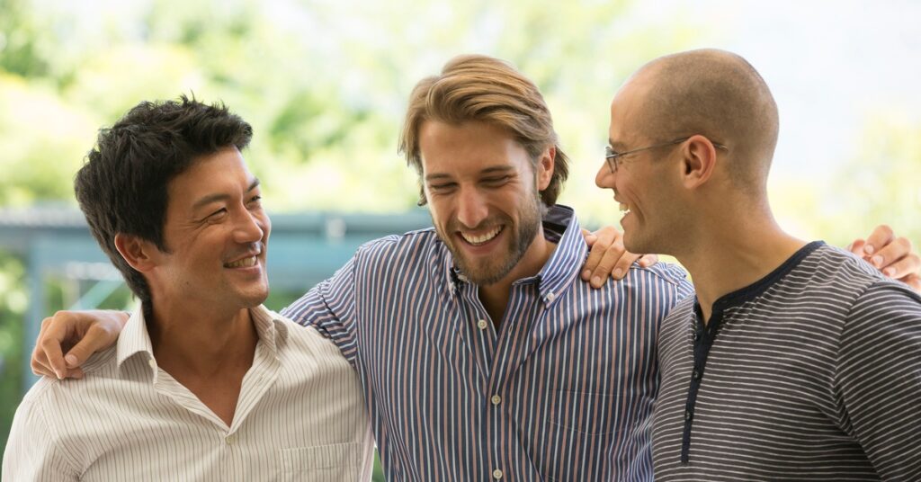 Three men stand outside together with their arms around each other's shoulders. They smile at each other.