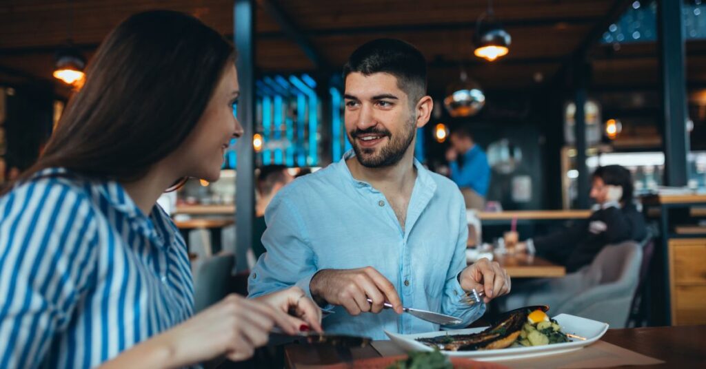 A man and woman look at each other as they converse in a restaurant. They both hold their cutlery over their plates of food.