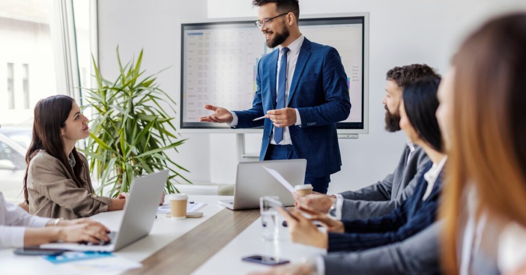 A businessman stands at a conference table and speaks in front of a TV screen. Other business professionals listen.