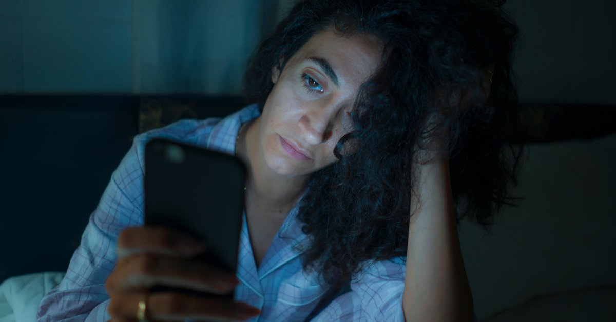 A woman in a dark room leans her head against her hand. She has a neutral expression as she looks at her cell phone.
