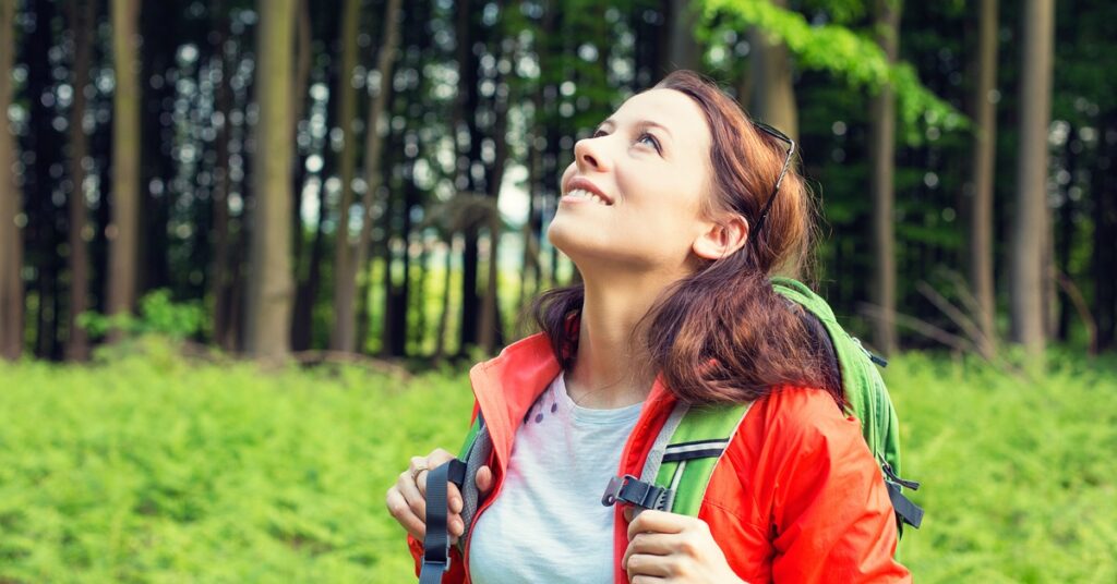 A woman in a wooded area looks up at the sky and smiles. She wears a backpack and hooks her thumbs on the straps.