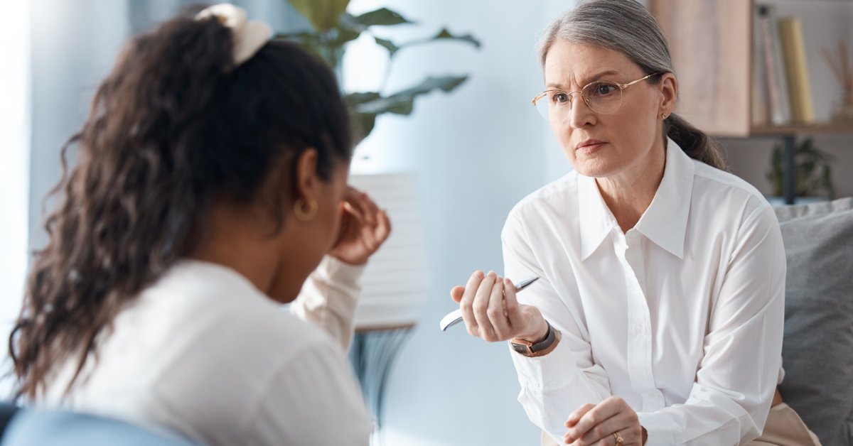 A therapist slightly frowns and leans toward her client. The client looks downward and touches her forehead.