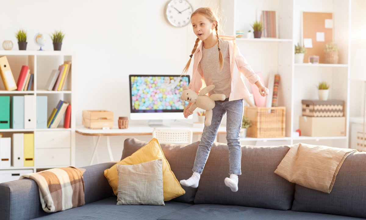 A little girl with pigtails is suspended mid-air as she jumps on a couch in a sunny room. She holds a white teddy bear.