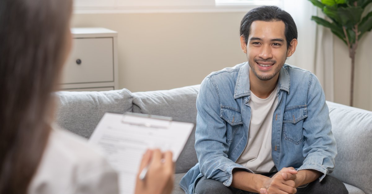 A man sitting on a couch smiles as a doctor writes notes on a clipboard. A window and a small tree are behind him.