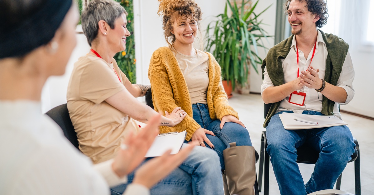 A diverse group of people sit in black chairs, smiling and talking to each other. A small table sits in the middle of them.