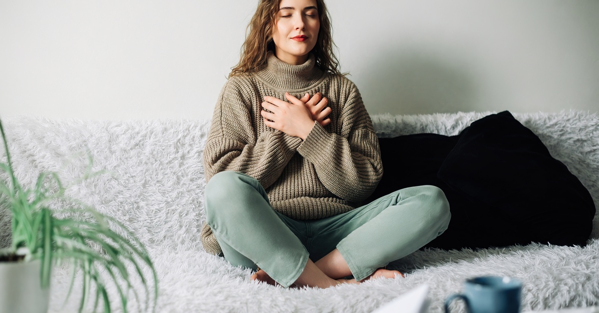 A woman wearing a brown sweater and green jeans sits on a couch, her legs crossed, and her eyes closed.