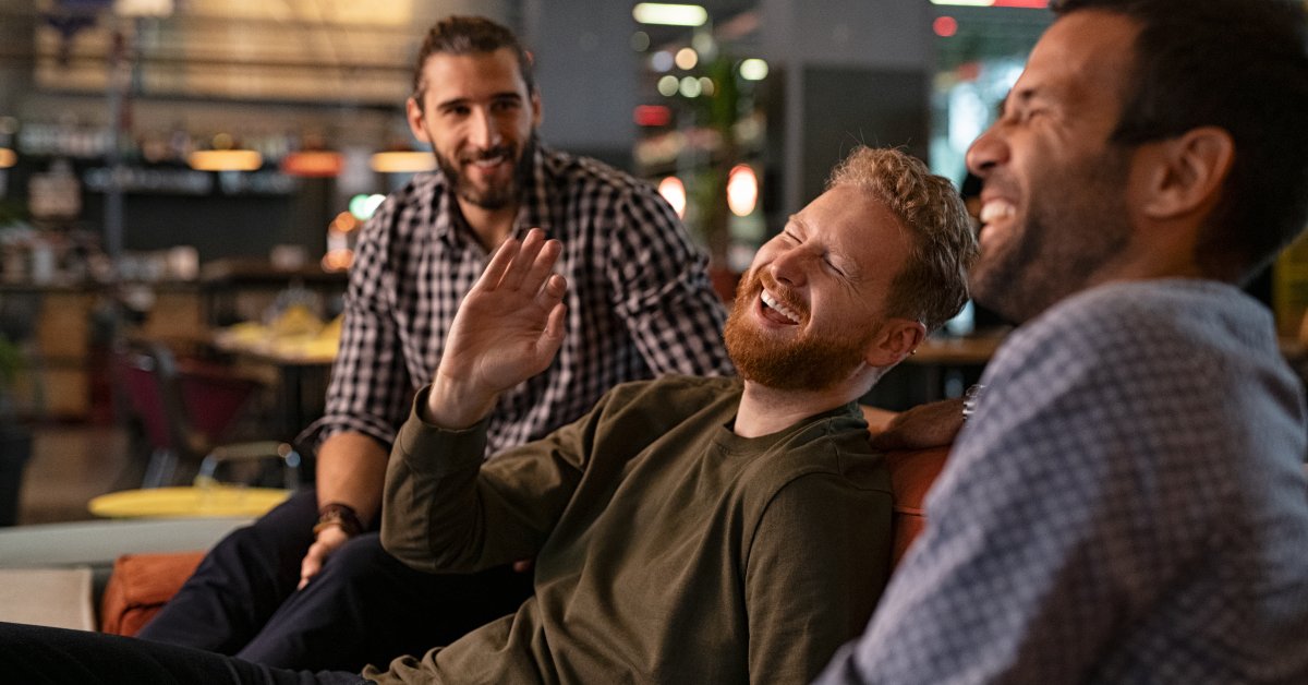 Three men sit and laugh together in a public space. The man in the middle tilts his head back and raises his hand.
