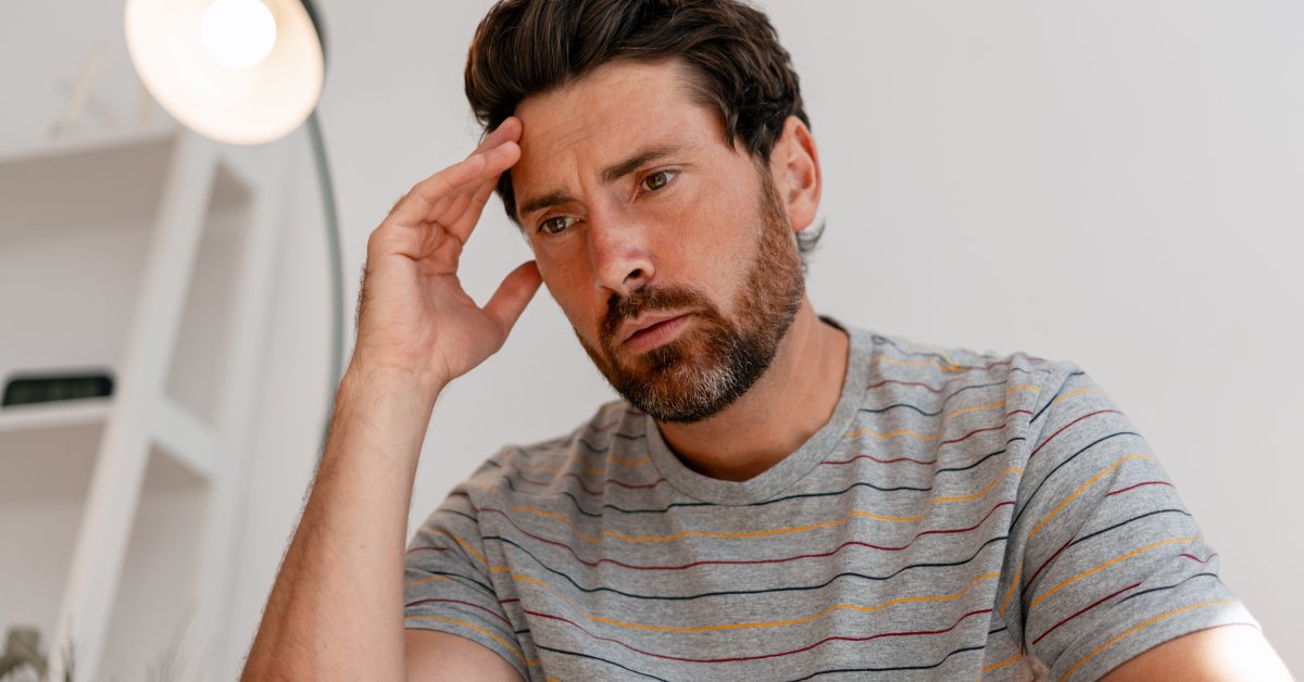 Upward view of a seated man who looks downward with a pensive expression. He gently touches his hand to his forehead.