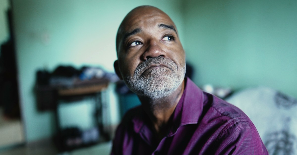 Side view of a seated man looking upwards to an unseen window. His lips are pressed shut, and his eyebrows are raised.