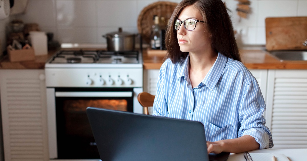 A woman wearing glasses sits at a kitchen table with her hands on a laptop keyboard. She stares into the distance.