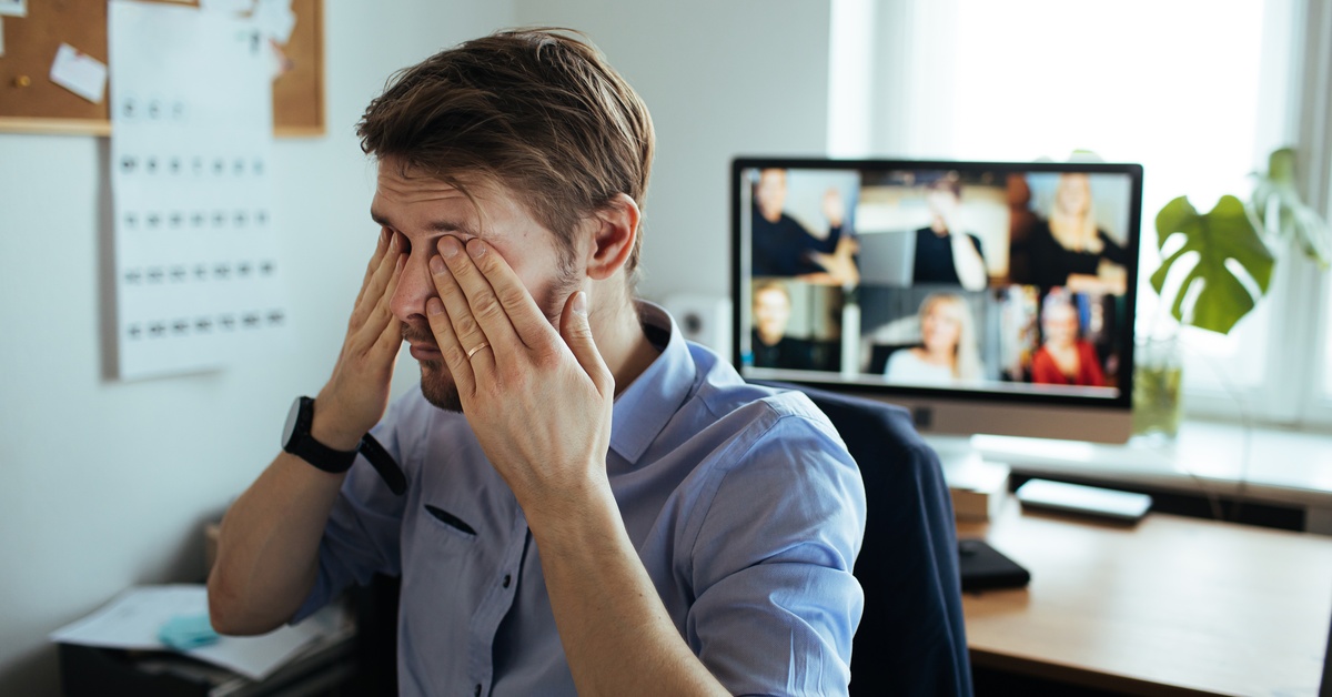 A man presses his hands against his closed eyes. Behind him, his blurred computer screen shows people in a video call.
