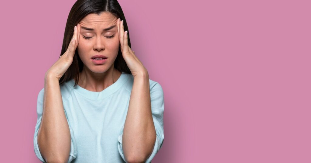 A woman with closed eyes presses her hands against either side of her head, causing her forehead to slightly furrow.