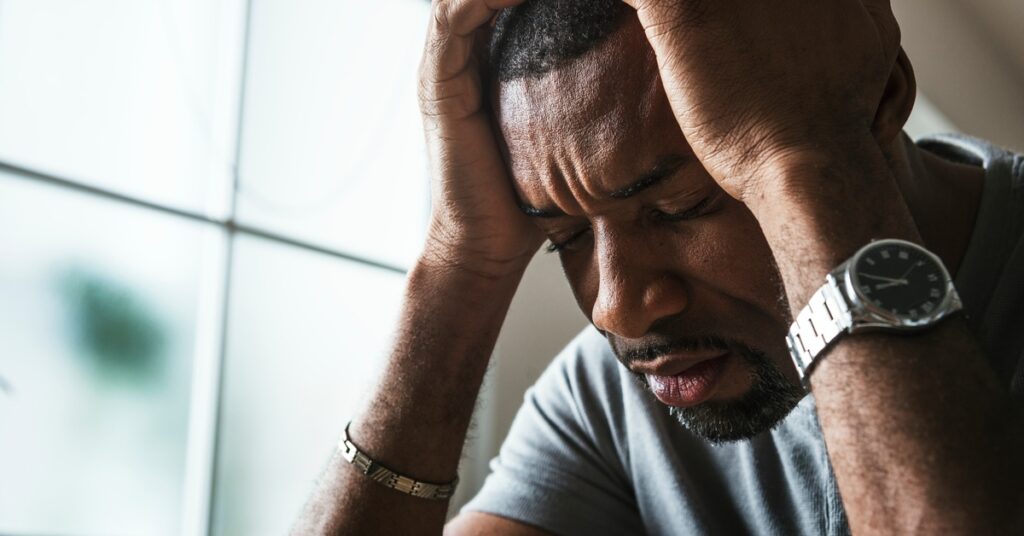 Close-up of a man near a frosted glass window. He presses his hands to his head and frowns with his eyes closed.
