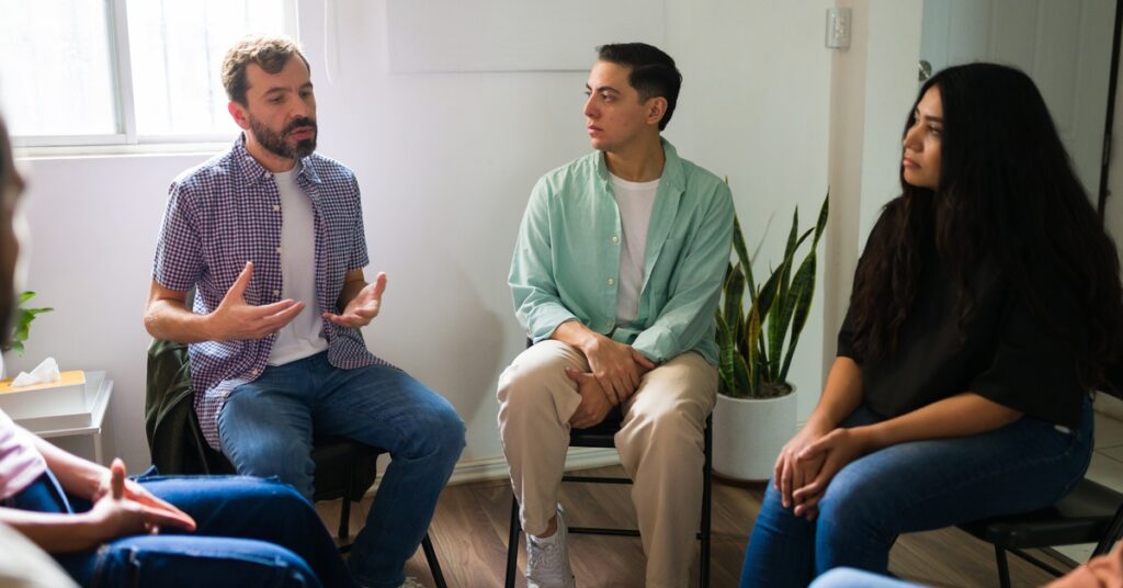 In a group of people seated in a circle, a man looks down at the ground as he talks. Those around him watch and listen.